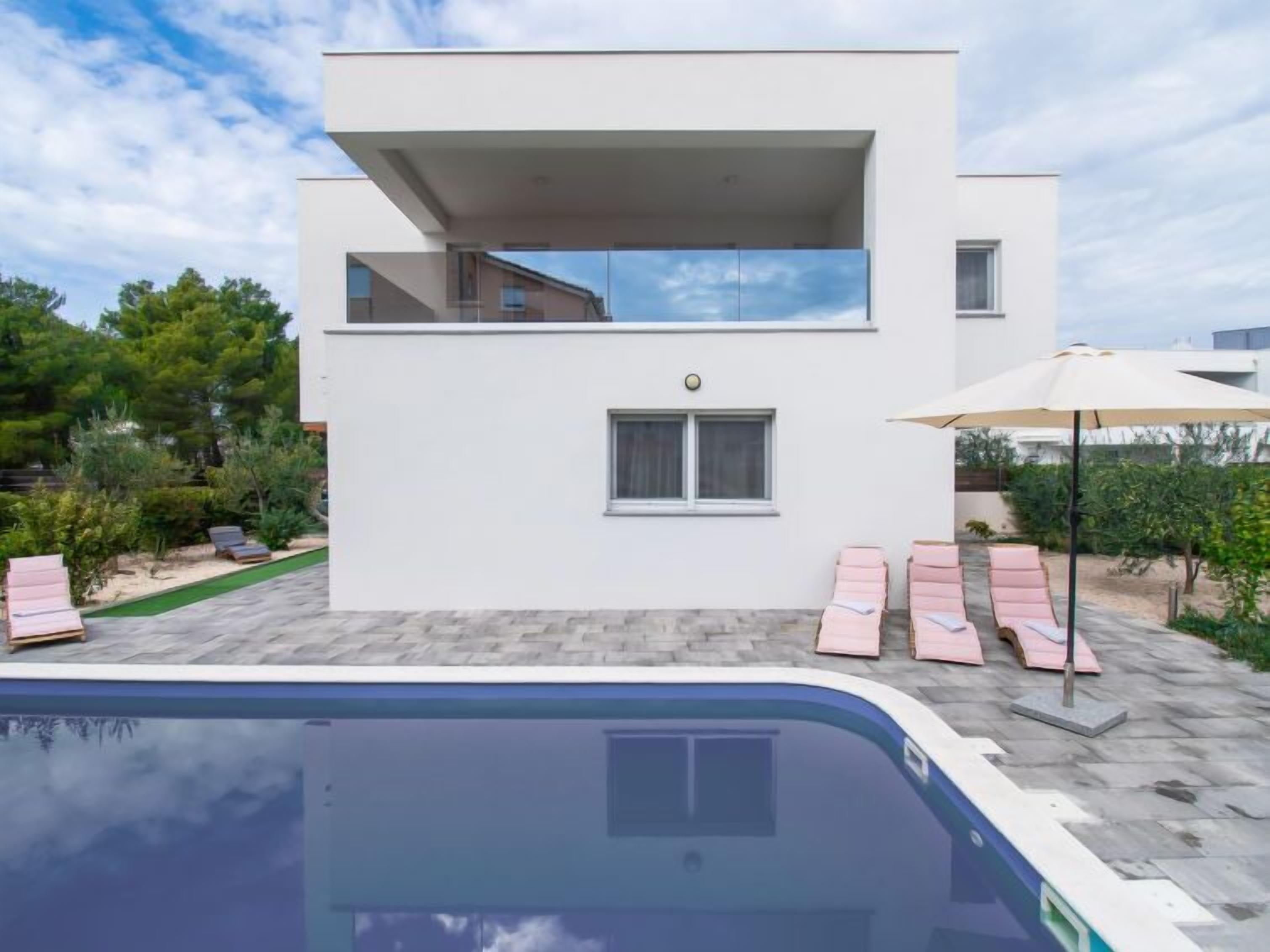 Une maison moderne à deux étages, dotée d’un balcon, avec une piscine en premier plan et un espace patio aménagé avec des chaises longues.