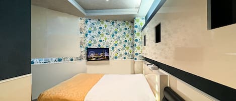 A compact hotel room with a bed, a small desk, a TV mounted on the wall, and a patterned wallpaper.