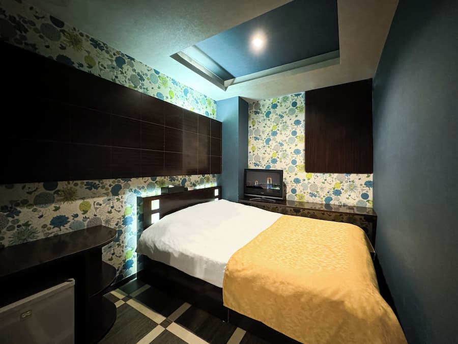 Quarto de hotel com cama, televisão, escrivaninha e um pequeno frigorífico.