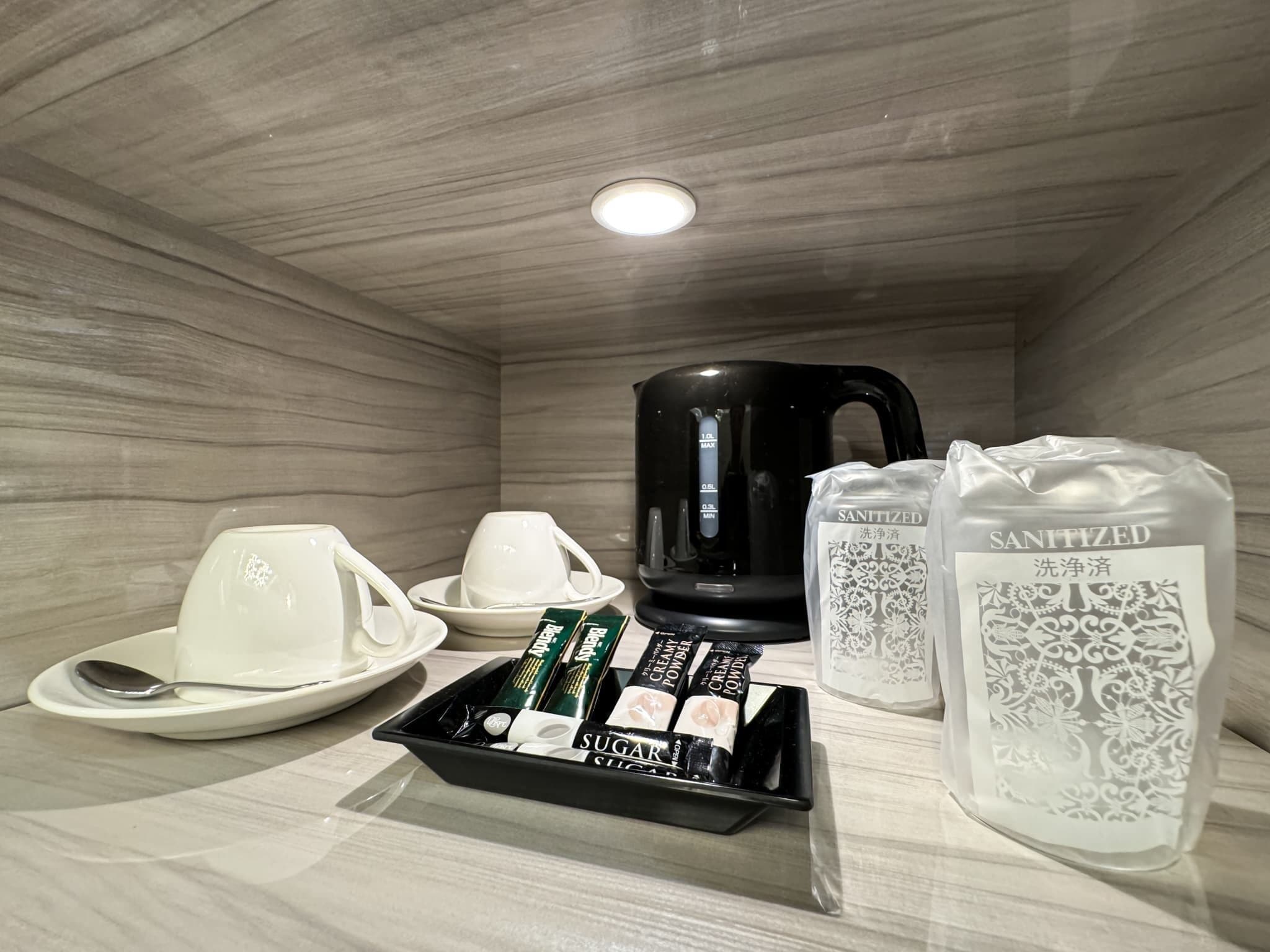 A hotel room with a black coffee maker, white cups, a tray with sugar packets, and a bag of hand sanitizer.