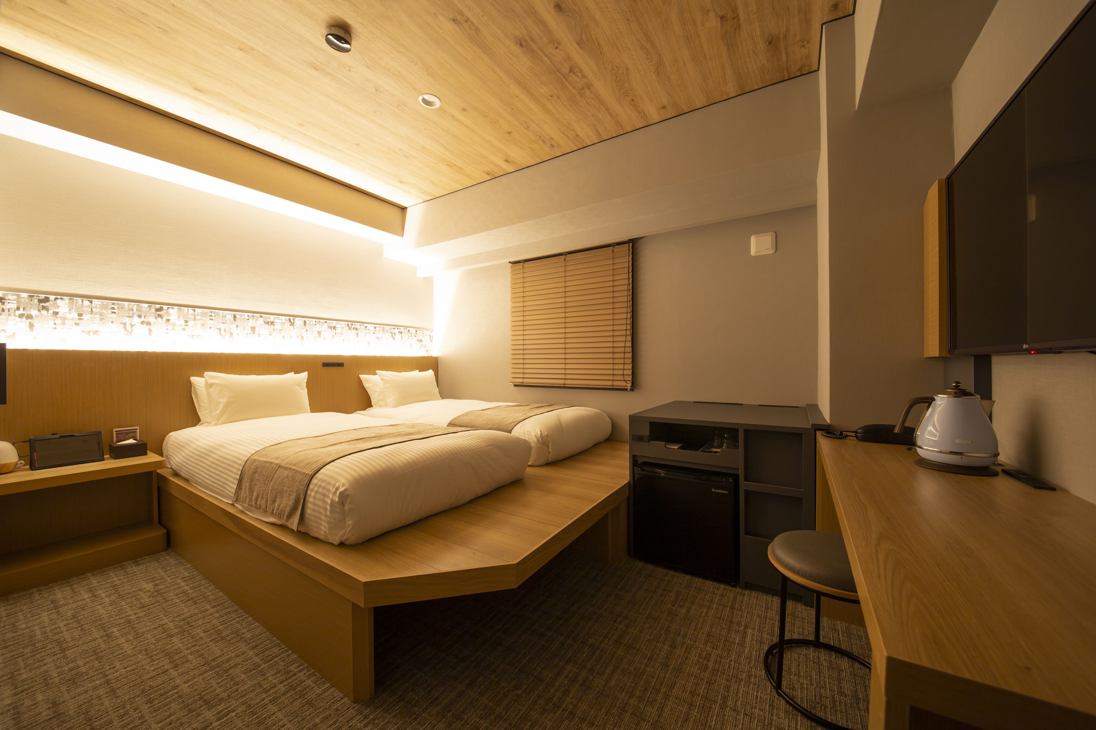 A modern hotel room with a bed, desk, and kitchenette.