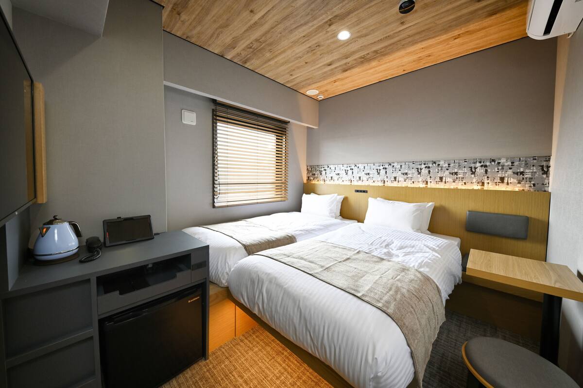 A hotel room with two beds, a desk, a chair, a small kitchenette, and a window with blinds.