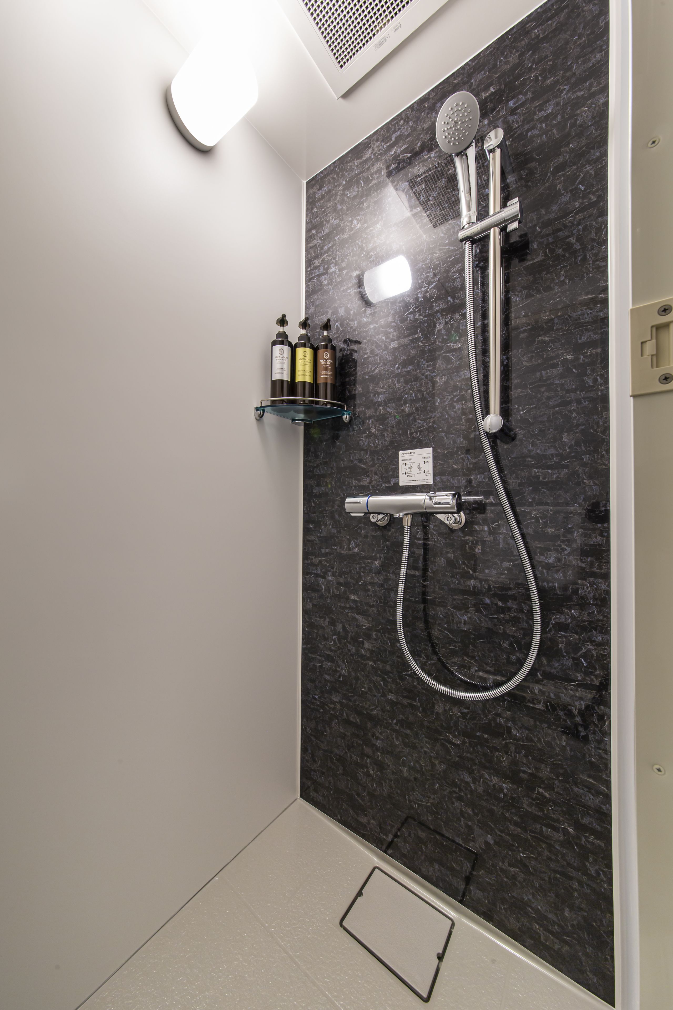 A modern shower with a dark marble wall, a handheld showerhead, a wall-mounted faucet, and a handheld shower hose. There are two bottles on a shelf above the shower.