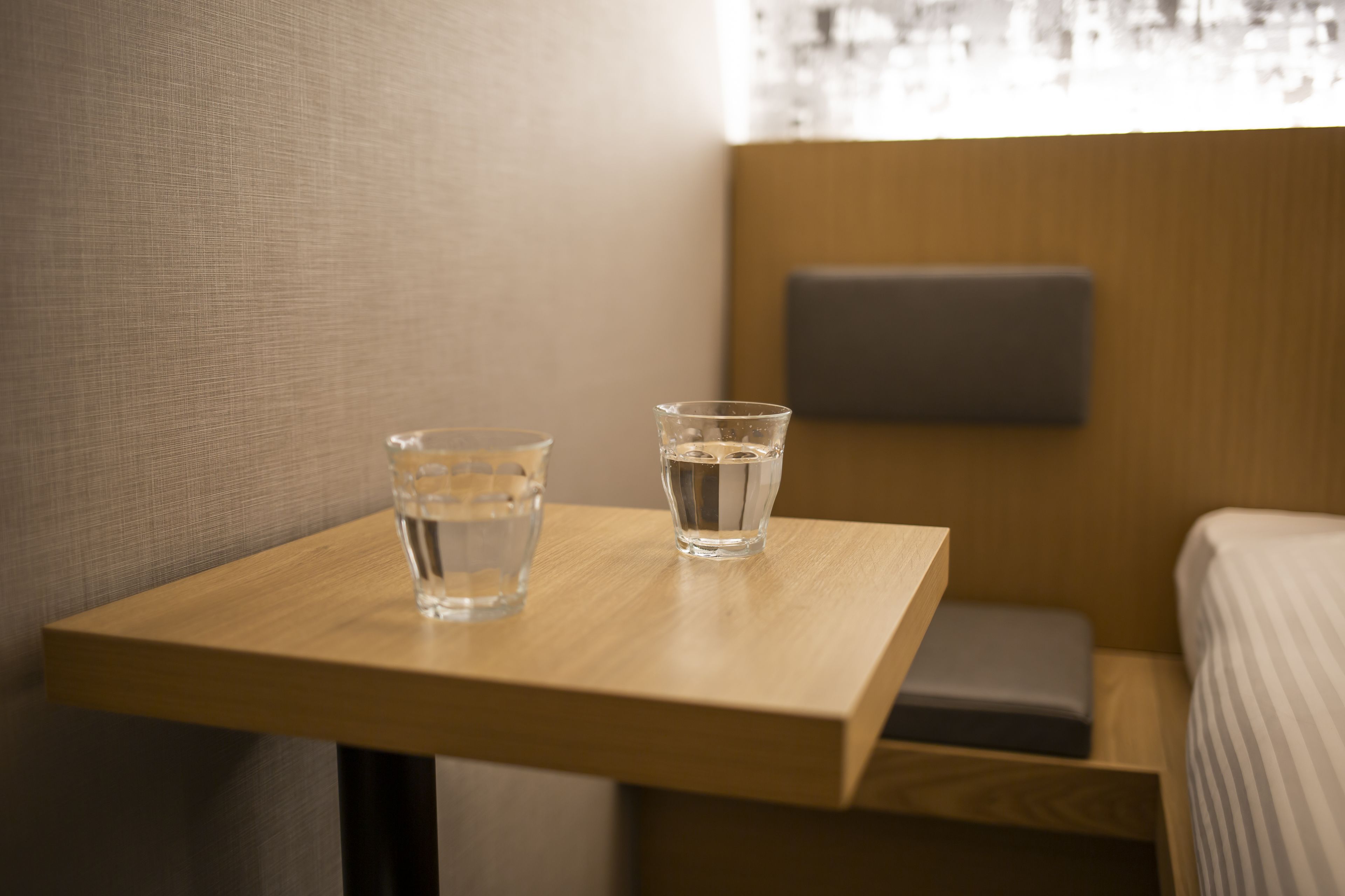 Ein kleiner Holztisch mit zwei Wassergläsern und einem grauen Kopfkissen auf einem Bett in einem Hotelzimmer.