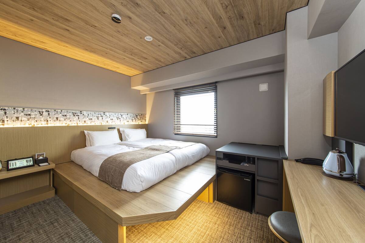 A modern hotel room with a wooden ceiling, a large bed with a wooden platform, a desk with a kettle, and a television.