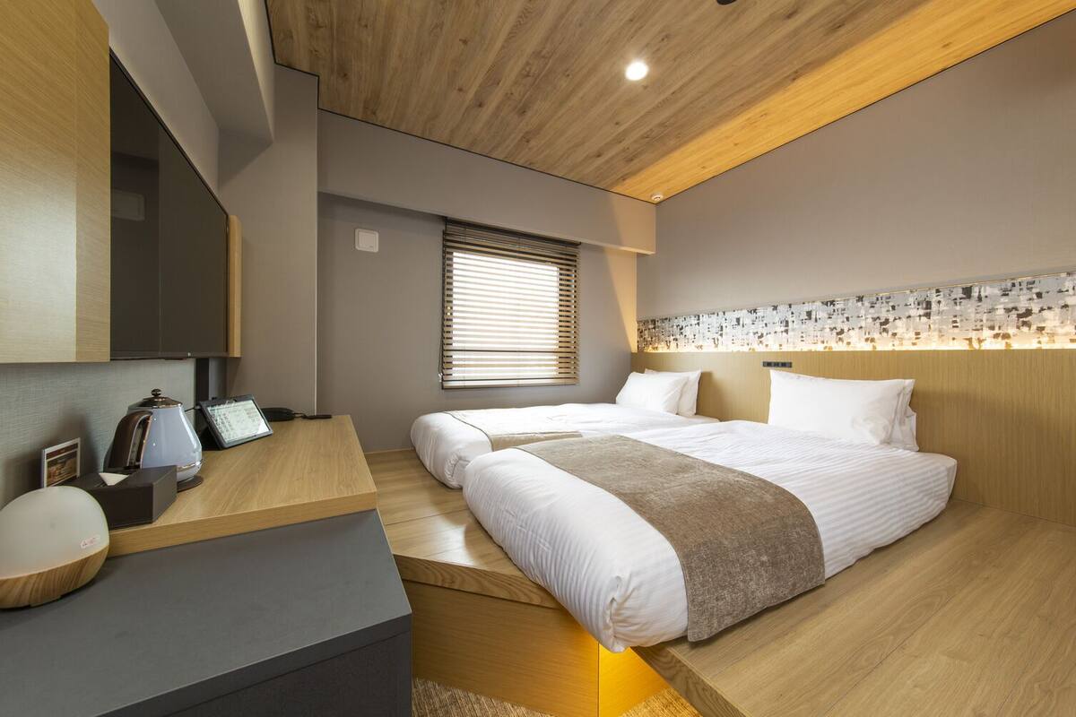 A modern hotel room with a wooden ceiling, a desk with a telephone and a coffee maker, a bed with white bedding, and a wall with a mosaic pattern.