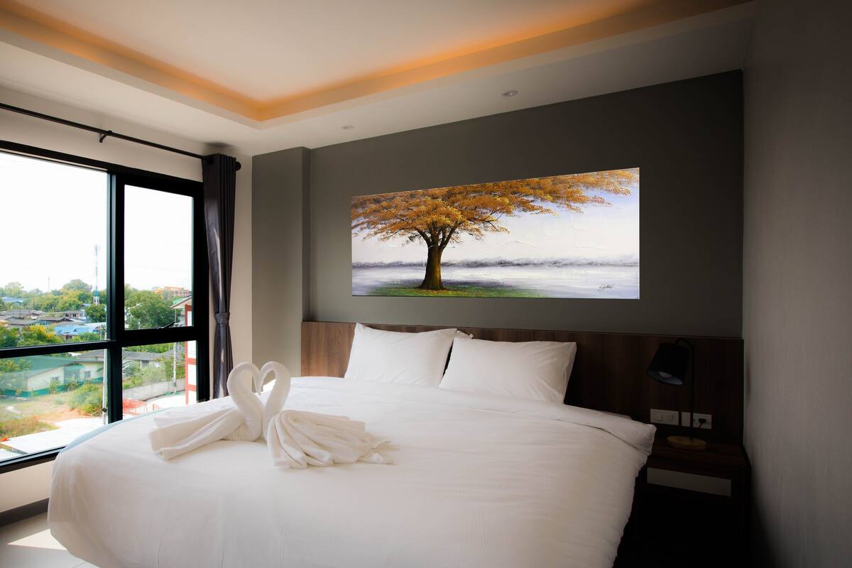A hotel room with a large bed, a painting of a tree, and a view of a cityscape through a large window.