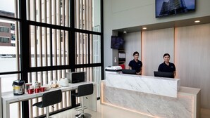 Un espace d’accueil d’hôtel avec un comptoir en marbre, deux réceptionnistes, un téléviseur diffusant des images d’un bâtiment et une fenêtre avec des stores.