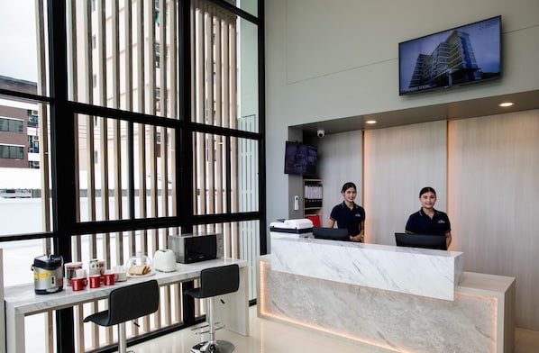 A hotel reception area with a marble counter, two receptionists, a TV displaying a building, and a window with blinds. - The Zleep Chonburi (Chonburi)