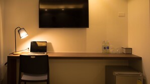 Une petite pièce faiblement éclairée, comprenant un bureau, une chaise, un ordinateur portable, un téléviseur, un mini-réfrigérateur et un climatiseur fixé au mur.