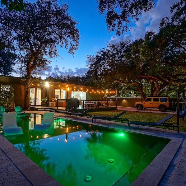 A swimming pool with lounge chairs, a covered outdoor seating area, and a vehicle parked nearby.