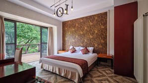 A modern hotel room with a large bed, a desk, and a balcony with a view of trees.