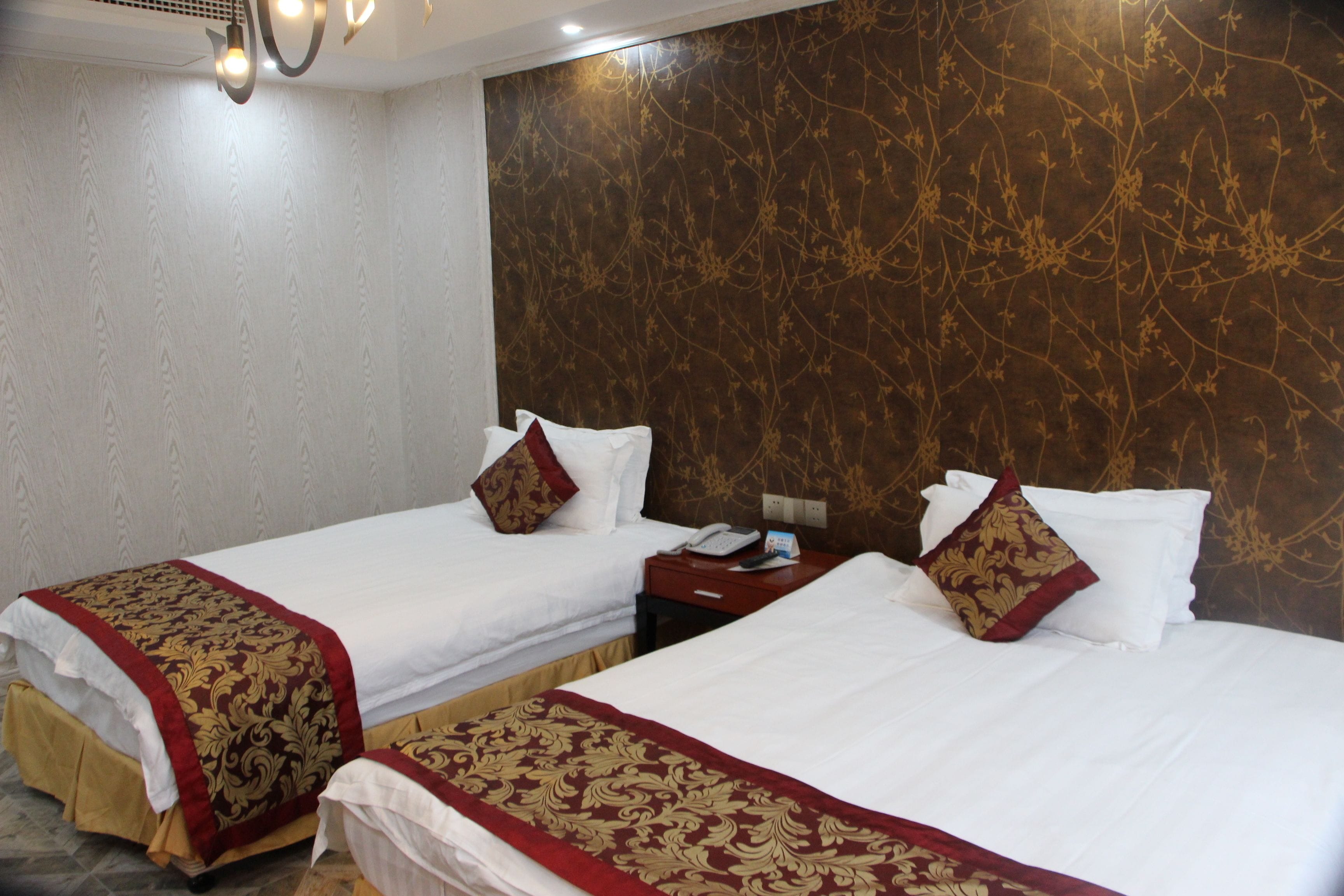 A hotel room with two single beds, each with a red and gold patterned bedspread, white pillows, and a wooden nightstand with a telephone.