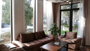 A spacious living room with a brown leather sofa, a matching armchair, and a wooden coffee table. Large windows offer a view of the outdoors.