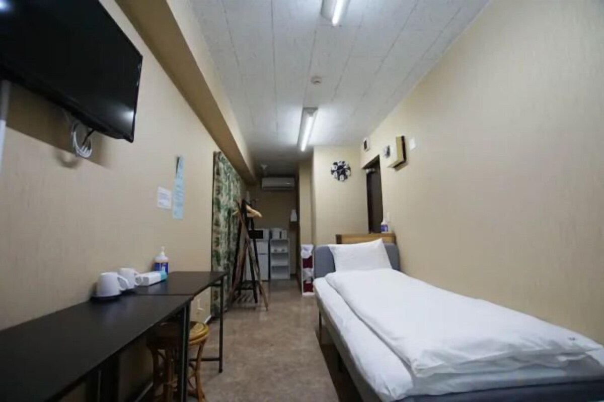 A narrow hotel room with a single bed, a desk, a chair, a television, and a door leading to another room.
