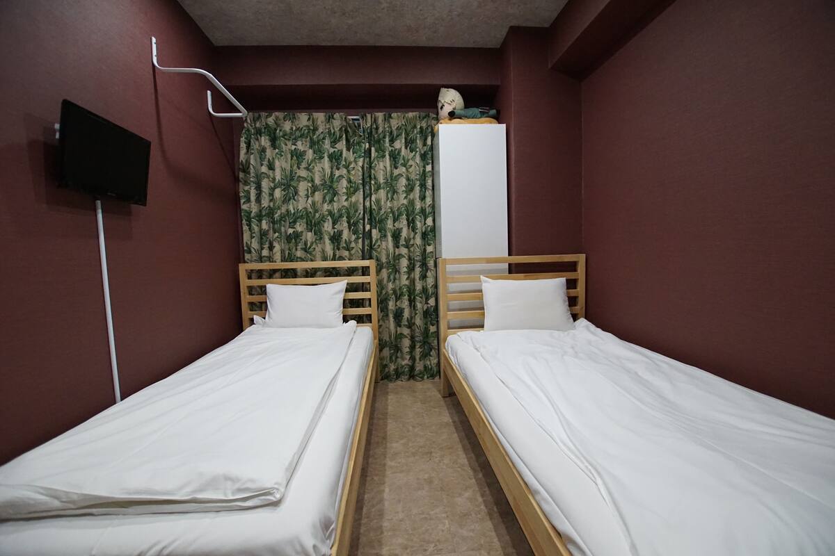 A room with two single beds, a television mounted on the wall, and a wardrobe in the background.