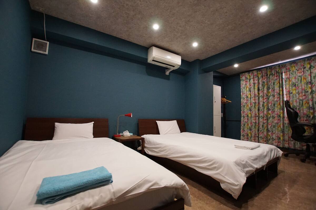 A hotel room with two single beds, a desk, a chair, an air conditioner, and a window with curtains.
