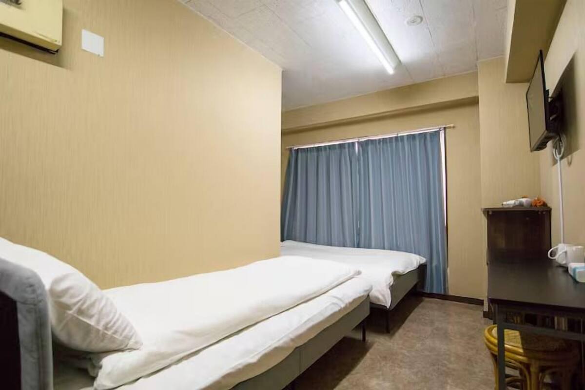 A room with two single beds, a small table with a chair, a television on the wall, and a window with curtains.