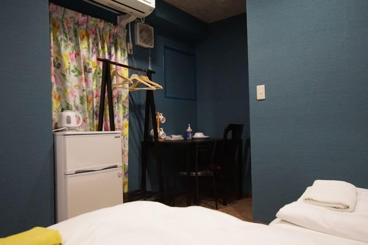 Ett kompakt hotellrum med en säng, ett litet skrivbord, en stol och en liten köksdel med ett kylskåp och en vattenkokare.