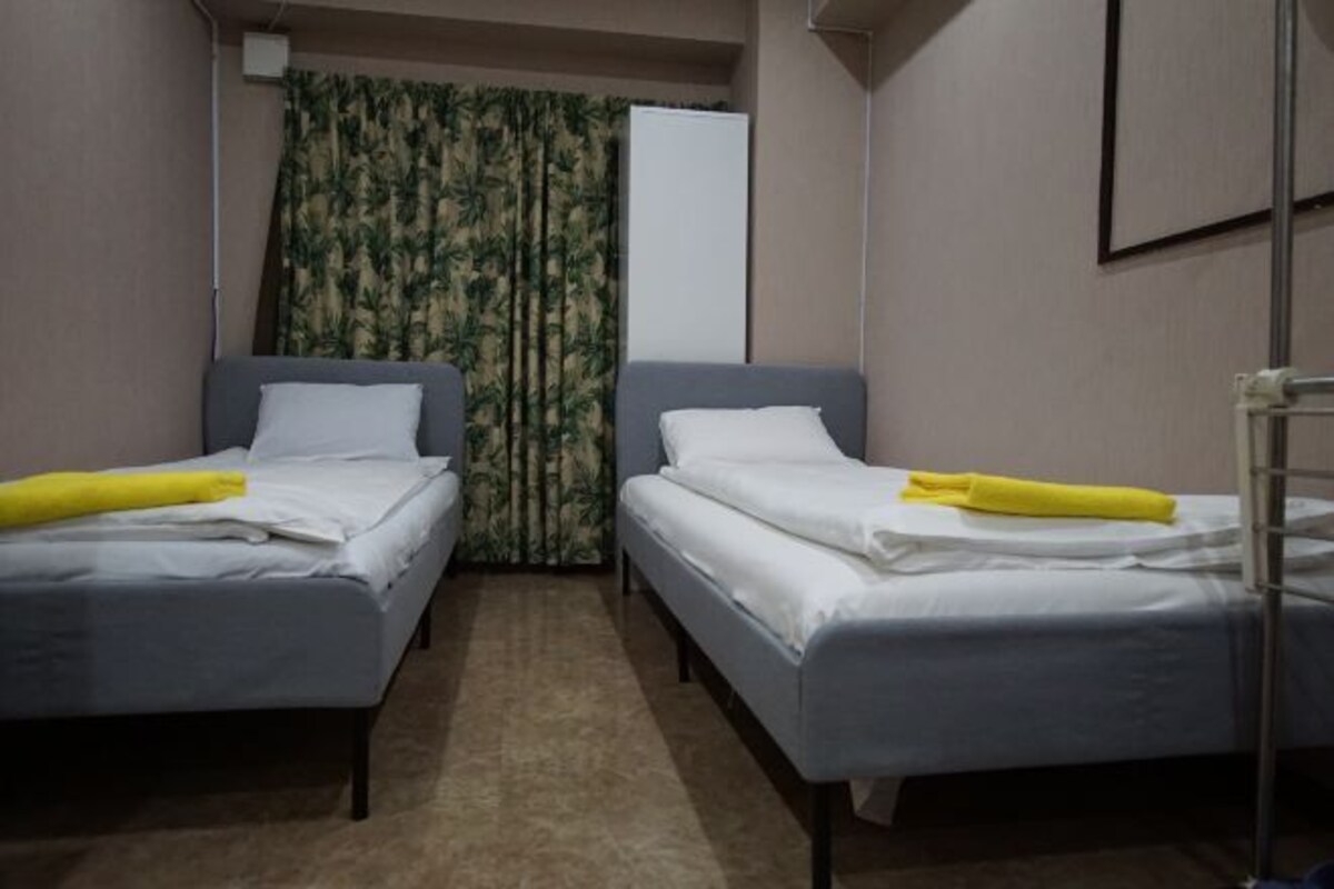 A room with two single beds, each with a white pillow and a yellow towel. The room has a carpeted floor and a curtain with a leaf pattern.