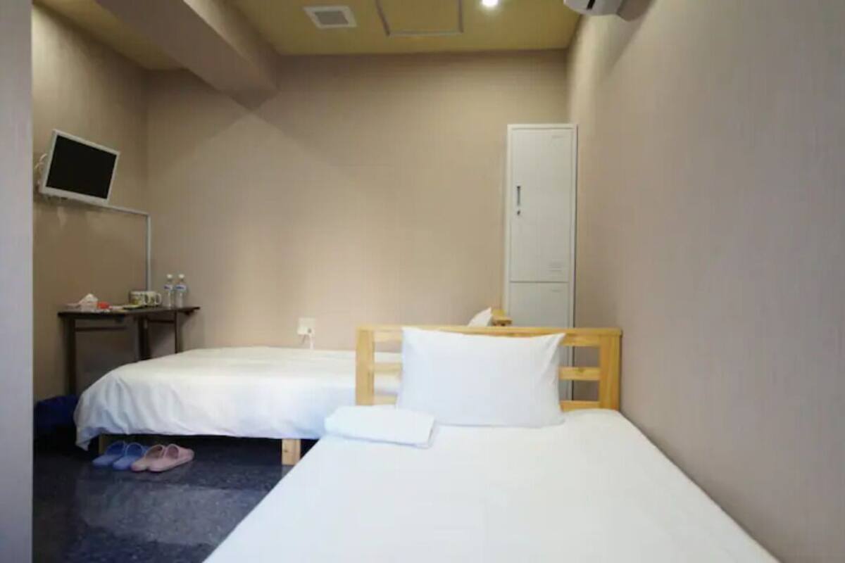 A small hotel room with two single beds, a desk with a computer, a wardrobe, and a TV mounted on the wall.