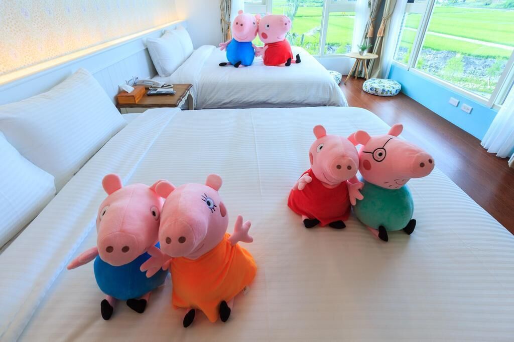 Habitació d'hotel amb dos llits, una finestra gran que mostra un paisatge verd i diversos peluixos amb forma de porc sobre els llits.