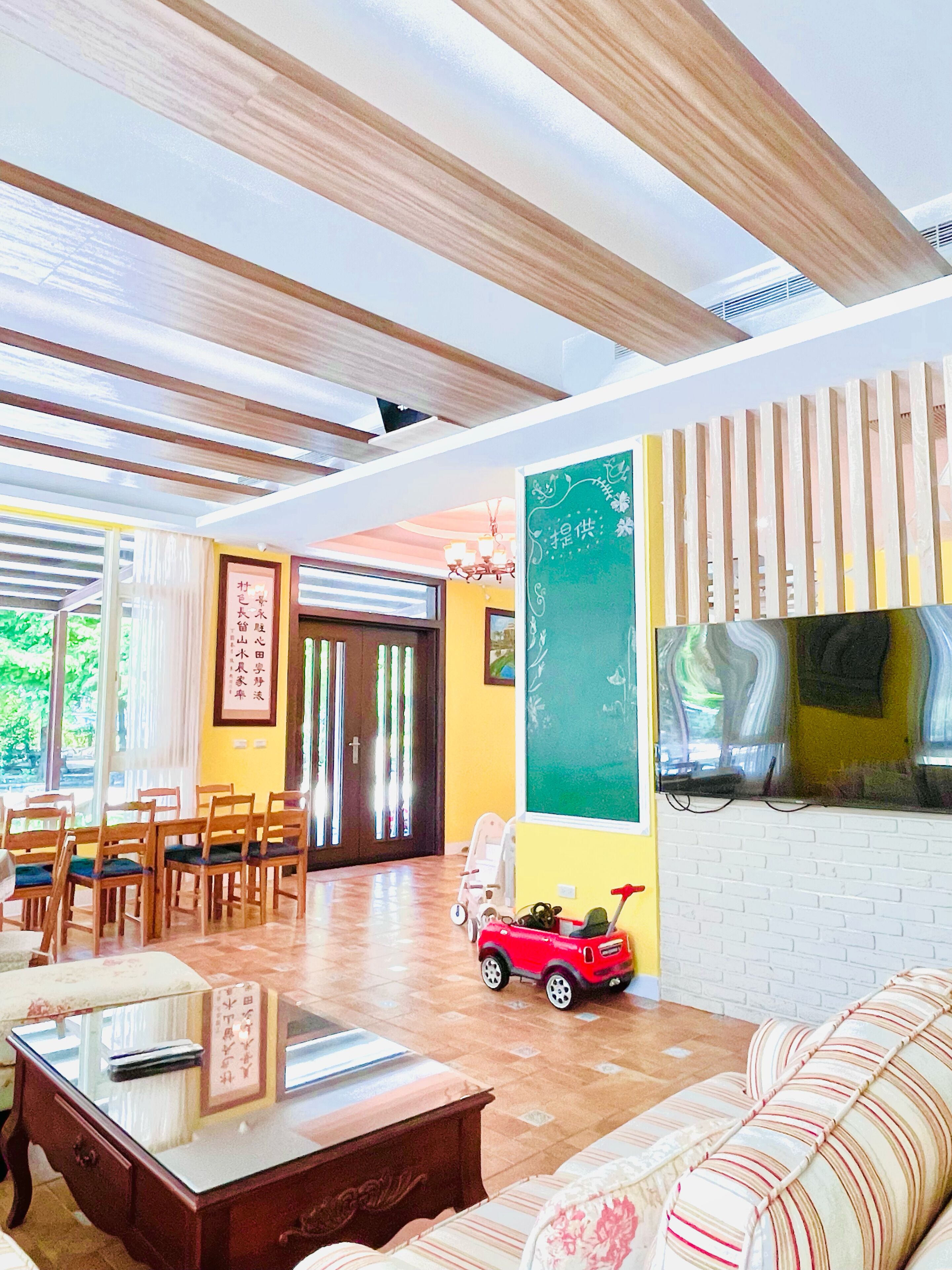 A room with a wooden ceiling, a sofa, a dining table with chairs, a small red toy car, and a chalkboard on the wall.