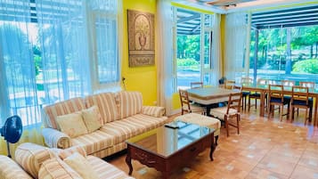 A spacious living area with a sofa set, a dining table with chairs, and a view of greenery outside.