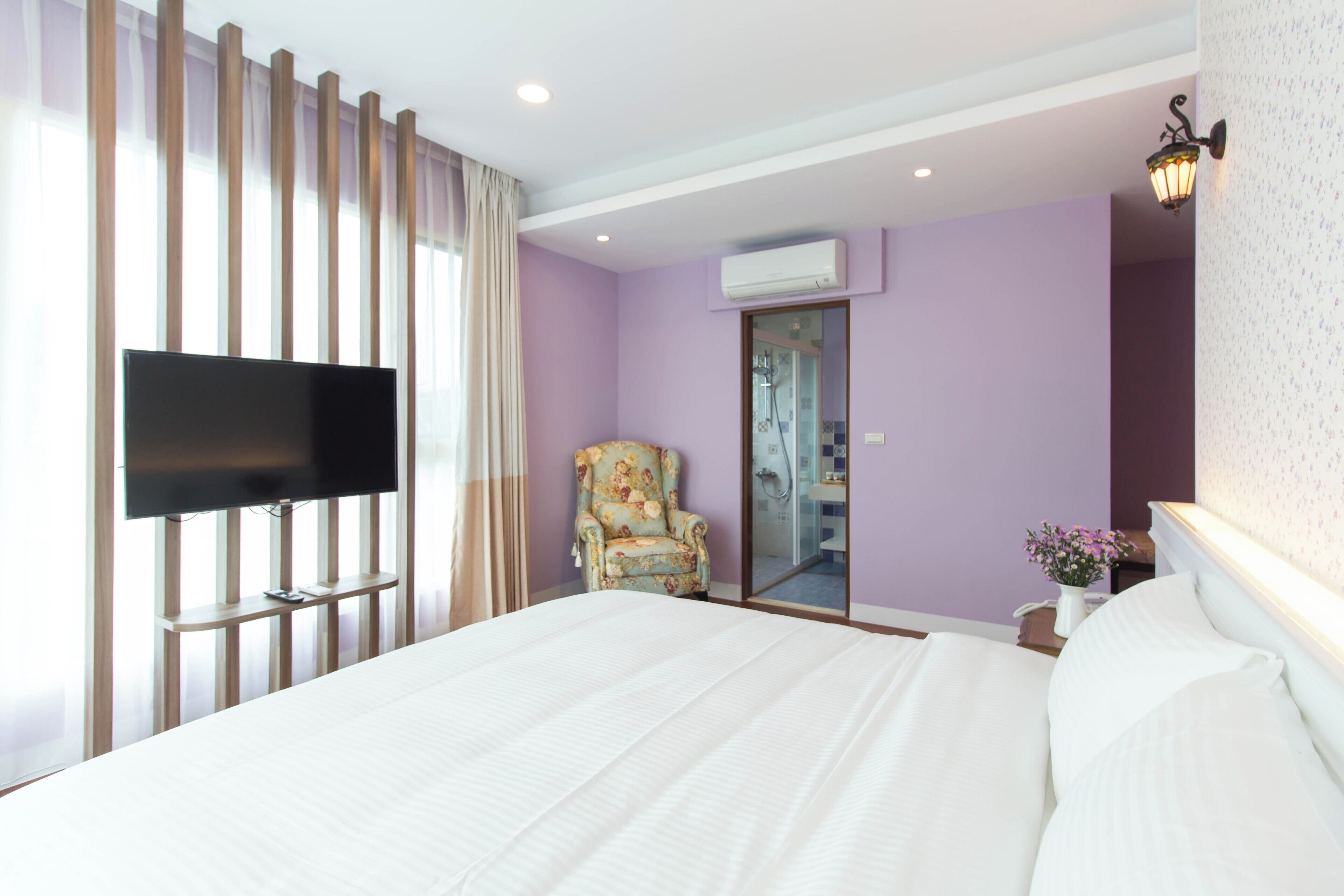 A hotel room with a large bed, a television mounted on a wooden panel, a floral armchair, and a bathroom visible through a glass door.