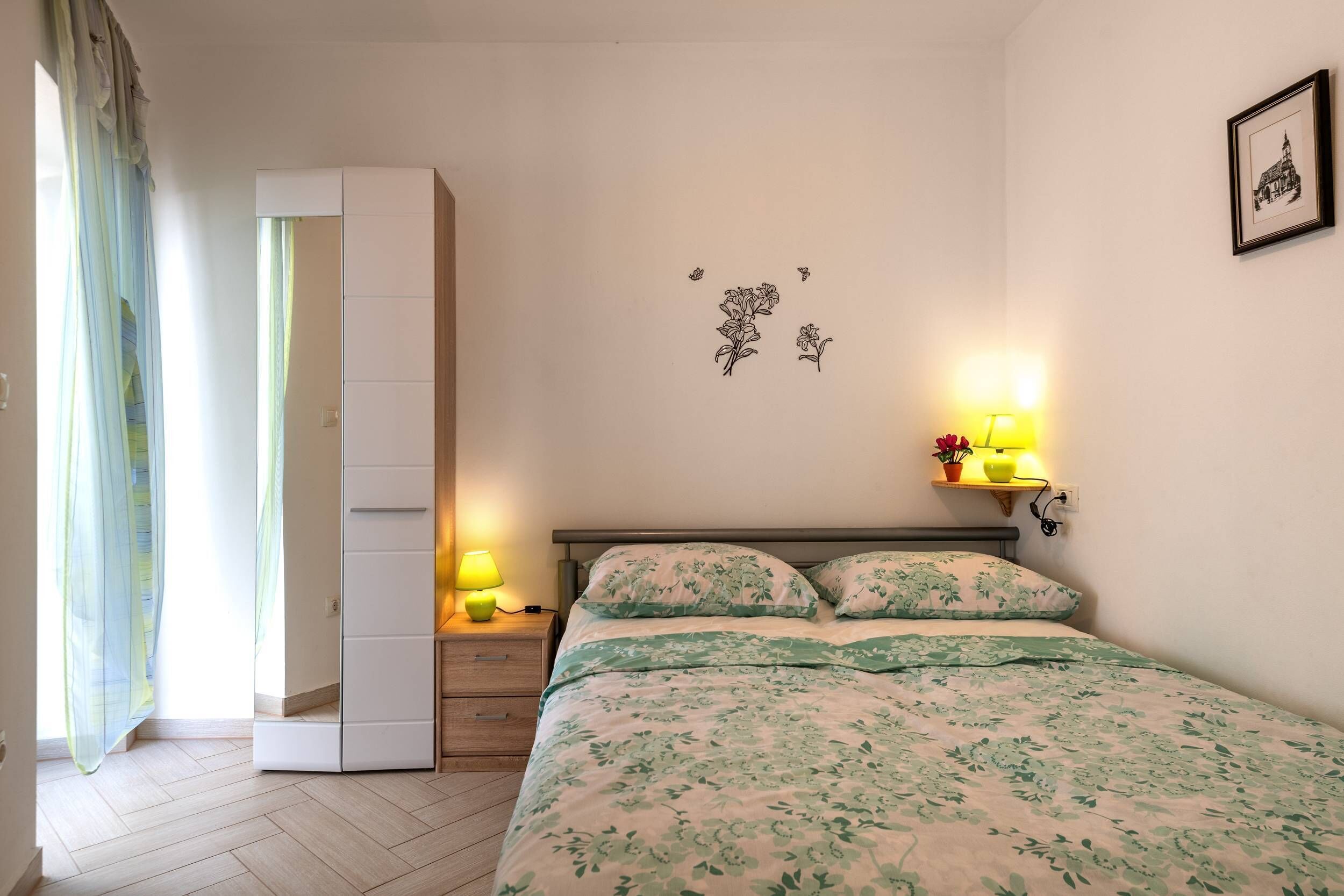 Um quarto com cama, mesa de cabeceira, guarda-roupa, luminária na parede e um quadro emoldurado.