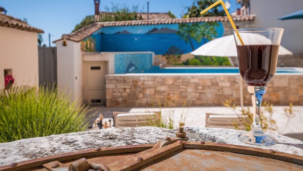 Ein Glas dunkles Getränk mit Trinkhalm auf einer rustikalen Metalloberfläche. Im Hintergrund ein Swimmingpool mit einem blauen Wandbild, umgeben von Grünpflanzen und einer Steinsäule.