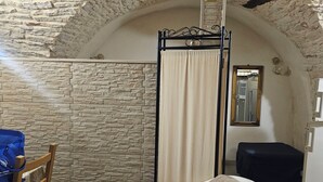 Ein Zimmer mit Steinwand, einem Bett, einem Sessel, einer Pendelleuchte und einem Spiegel.