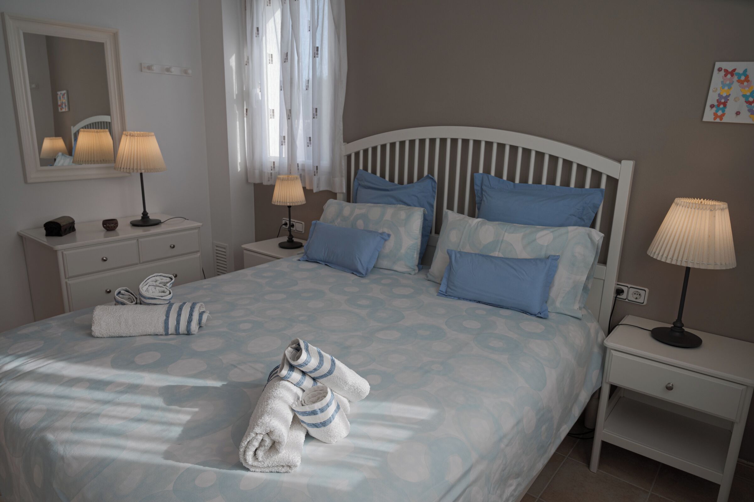Cama bem arrumada com roupa de cama azul e branca, mesa de cabeceira com abajur, espelho e pequena mesa de cabeceira com abajur.
