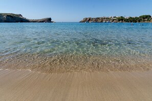 Ein Strand mit klarem Wasser, sandigem Ufer und felsigen Klippen im Hintergrund.