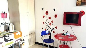 A room with a white cabinet, a table with a colorful tablecloth, two blue chairs, a red table, a red TV, and a wall decoration with flowers and birds. - Yi Xin Apartment - Hostel (Xi'an)