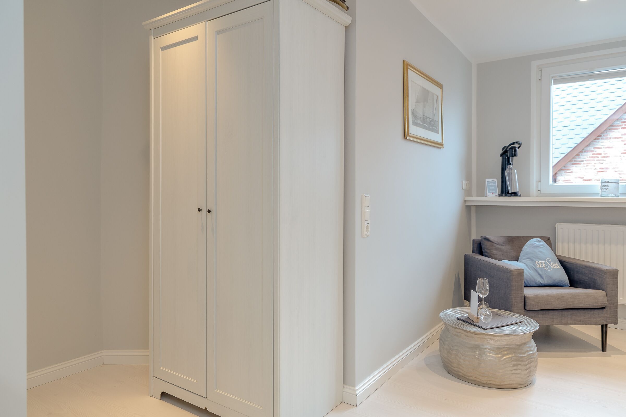 Une pièce avec une armoire blanche, un fauteuil gris avec un coussin, une table ronde avec un vase, et un tableau encadré accroché au mur.