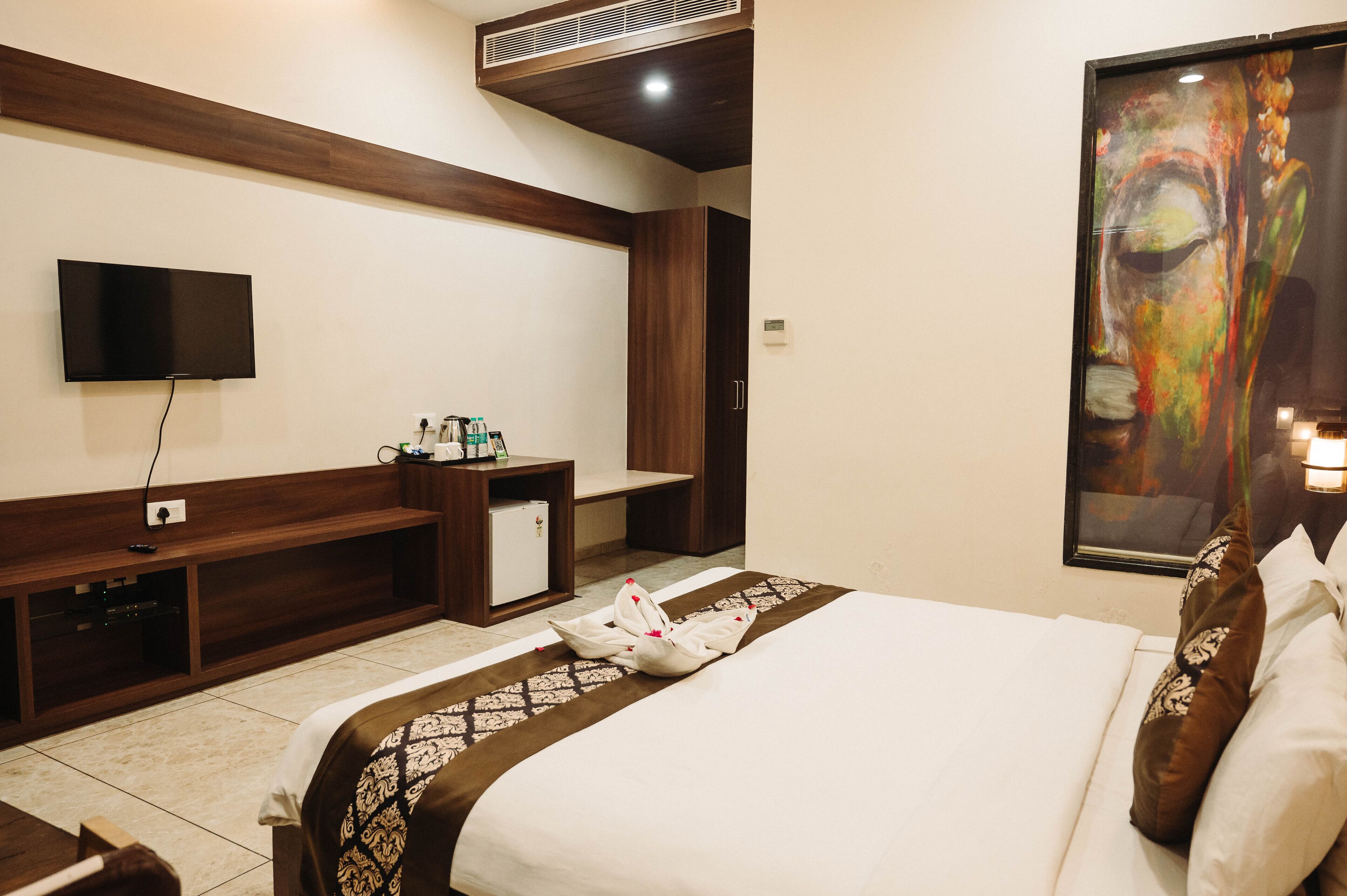 A hotel room with a bed, a flat-screen TV mounted on the wall, a small desk with a refrigerator, and a painting on the wall.