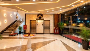 Un vestíbulo de hotel con una imponente escalera, ascensores, un mostrador de recepción y una gran planta en maceta.