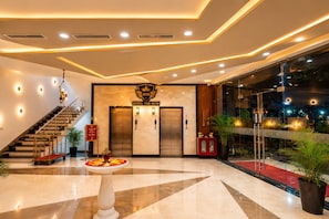 Un vestíbulo de hotel con una imponente escalera, ascensores, un mostrador de recepción y una gran planta en maceta.