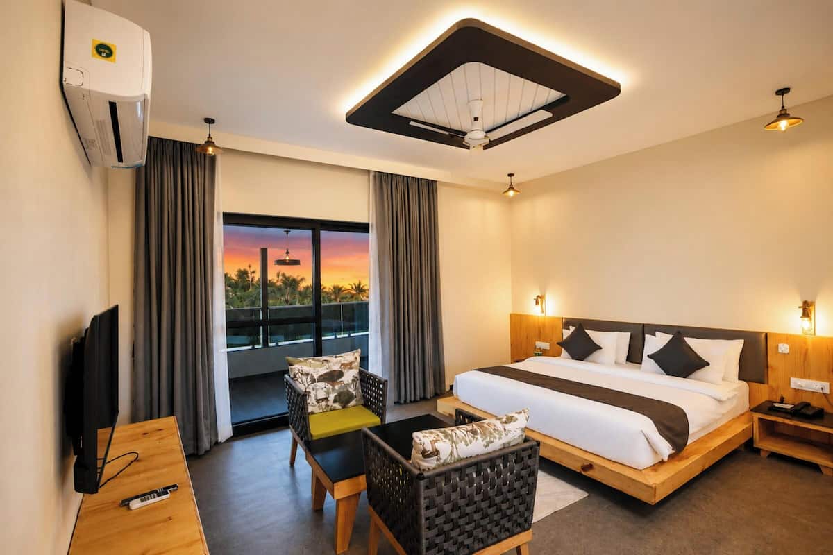 Habitación de hotel con cama, escritorio con televisor, silla, sofá, balcón con cortinas y ventilador de techo.