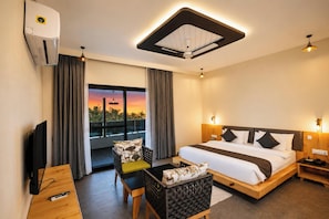 Habitación de hotel con cama, escritorio con televisor, silla, sofá, balcón con cortinas y ventilador de techo.