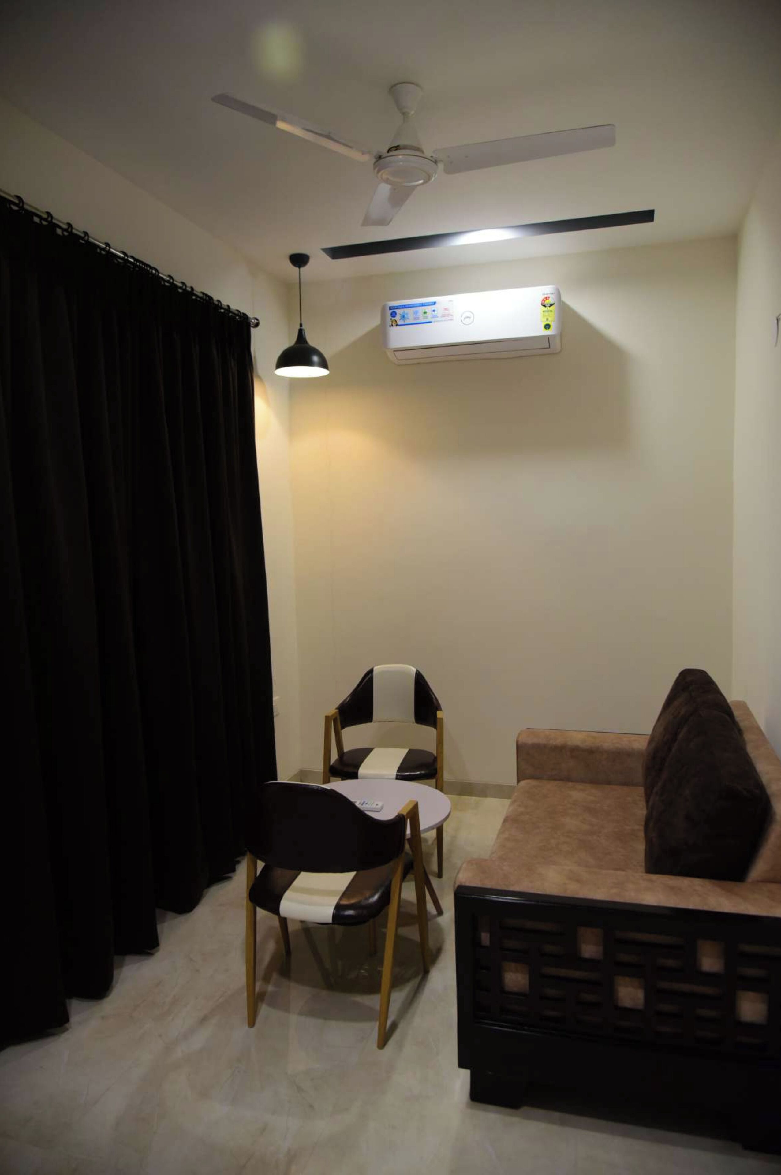 Una stanza con ventilatore a soffitto, condizionatore d'aria, una sedia bianco e nero, un divano marrone e una tenda nera.