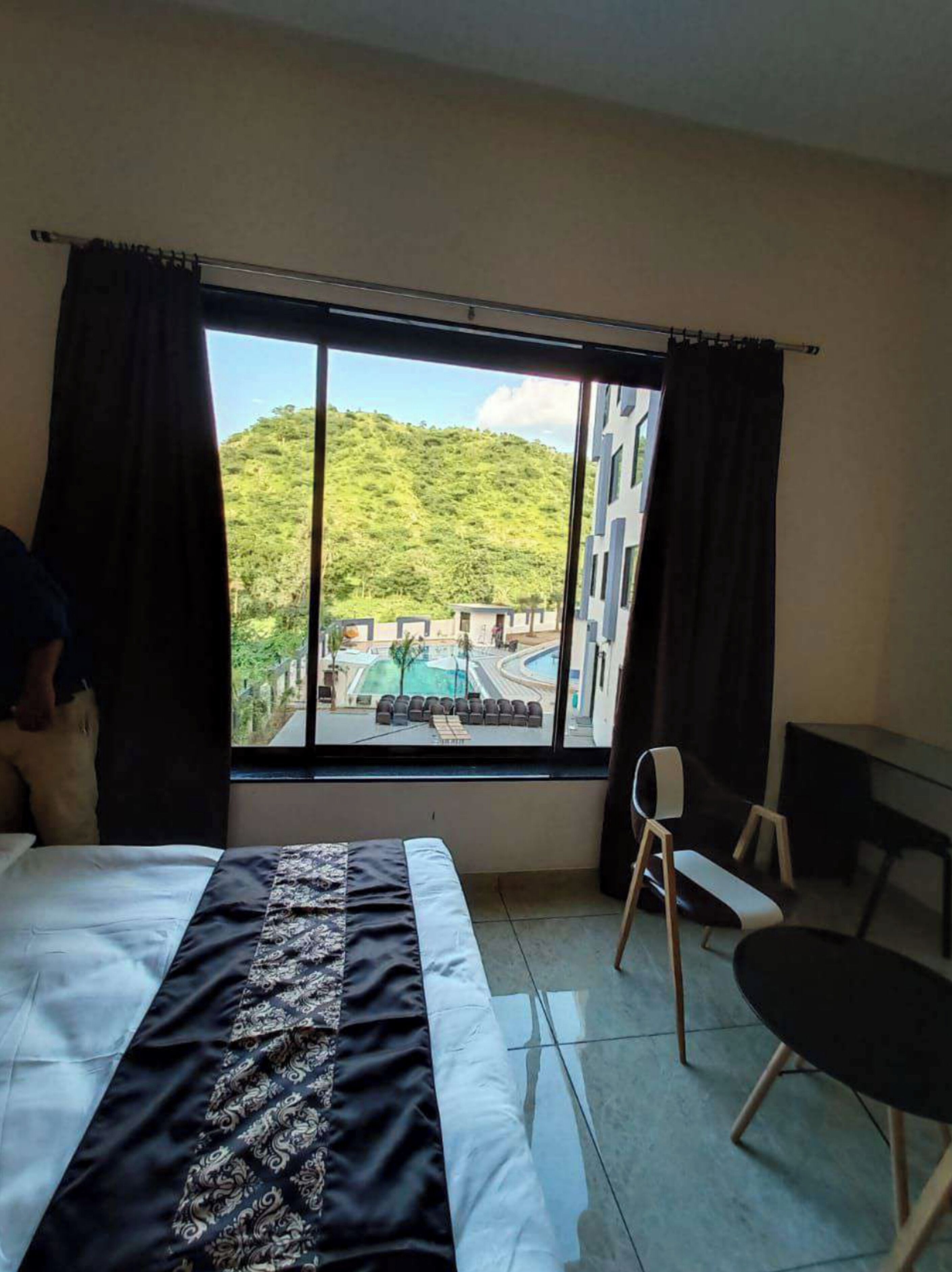 Habitación de hotel con cama, una silla, una mesita y un ventanal grande que ofrece vistas a una piscina y follaje.