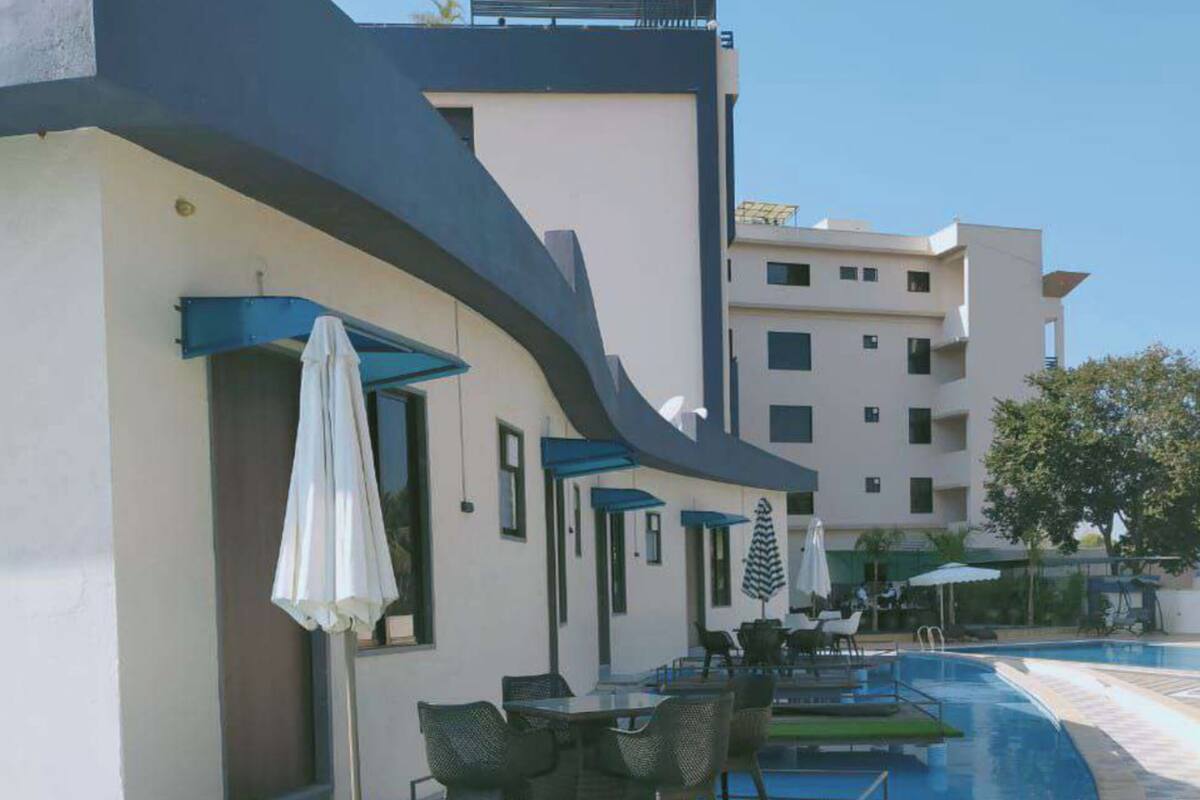 Zona junto a la piscina con sillones de descanso y sombrillas, piscina con azulejos azules y un edificio con fachada curva.