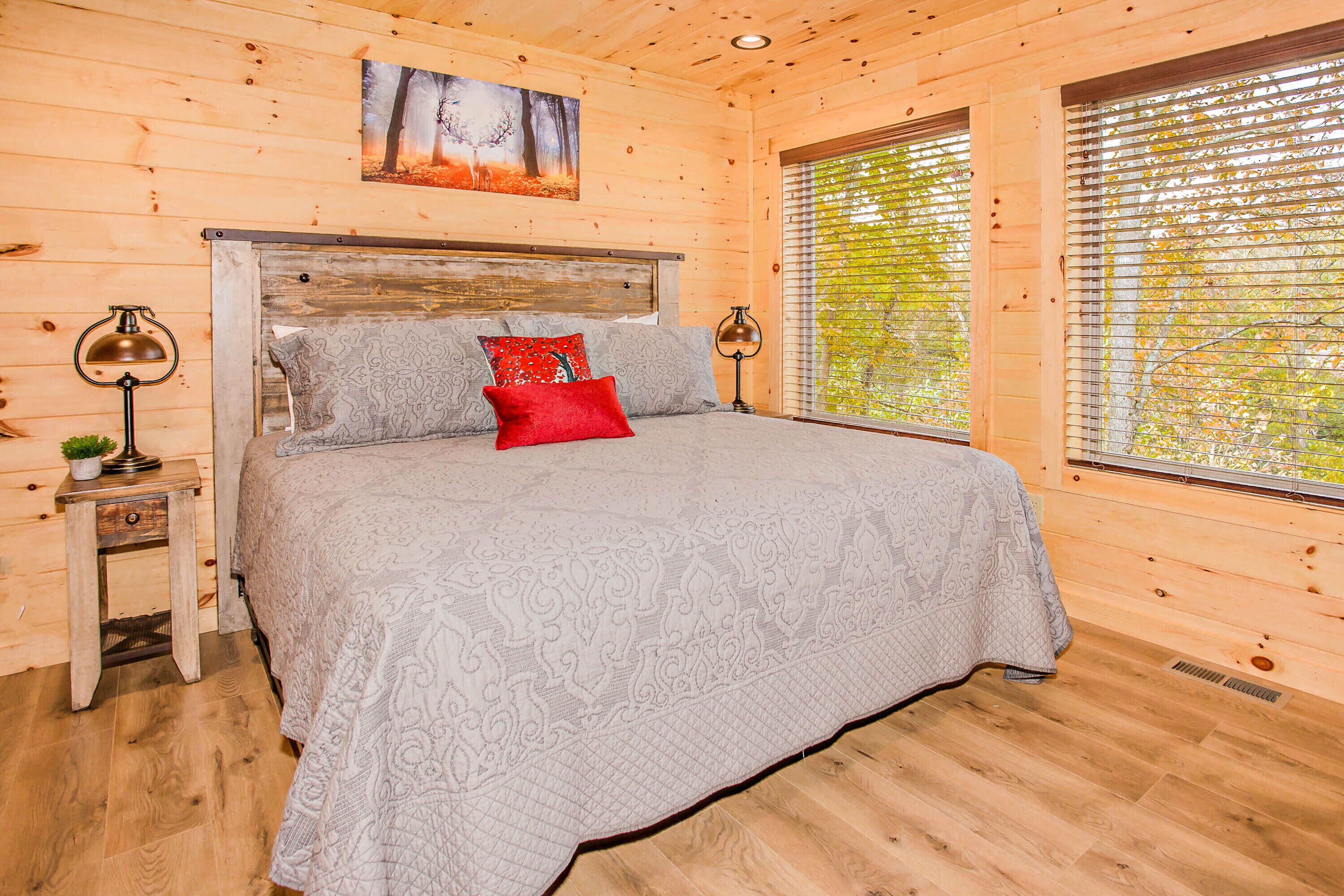 A wooden cabin bedroom with a large bed, two bedside lamps, a nightstand, and a window with blinds.