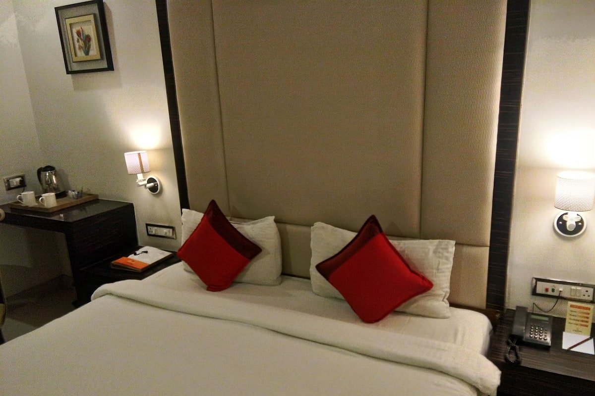 Habitación de hotel con una cama grande, dos almohadas rojas, una mesita de noche con lámpara, un teléfono y un pequeño escritorio con cafetera y un libro.