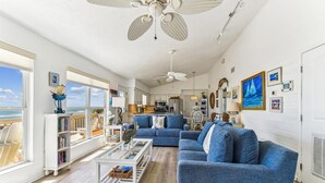 A spacious living room with a large blue sofa, a glass coffee table, and a view of the ocean through large windows. - SHARE A HEAVENLY VACATION with your family, friends, and pets! (St. George Island)