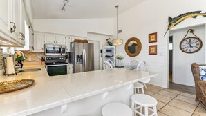 A modern kitchen with white cabinetry, stainless steel appliances, a large island with stools, and a dining area with a wicker chair and a wall clock. - SHARE A HEAVENLY VACATION with your family, friends, and pets! (St. George Island)