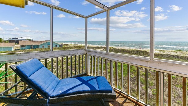 A balcony with a blue cushioned chair, overlooking a beach with waves and buildings in the distance. - SHARE A HEAVENLY VACATION with your family, friends, and pets! (St. George Island)