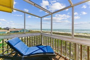 A balcony with a blue cushioned chair, overlooking a beach with waves and buildings in the distance. - SHARE A HEAVENLY VACATION with your family, friends, and pets! (St. George Island)